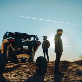 Can-Am SSV von hinten stehend in Wüste, zwei Fahrer stehen seitlich davor
